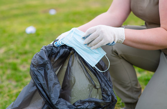 The Hands Of An Environmentalist Put A Used Medical Mask In A Black Bag. A Volunteer Cleans Nature From The Effects Of The Coronavirus Pandemic. Ecology, Environmental Protection Concept.