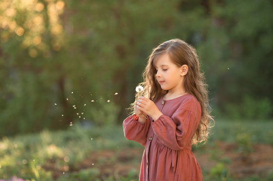 A girl in a pink dress and with her hair down stands against the background of greenery and sunset and blows on a dandelion.