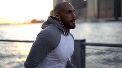 Slow motion of male runner in trendy sportswear jogging during sunset time on urban setting at metropolitan bund listening playlist in earphones for getting sport motivation for goal
