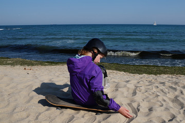 girl in a lilac jacket and a protective helmet sits on a skateboard on the beach