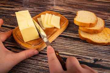 Fototapeta na wymiar Someone cuts some butter with a knife from a piece of butter in a wooden butter dish, slices of cheese and fresh wheat toast on a wooden background. Close up.