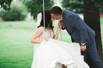 Romantic, fairytale, happy newlywed couple kissing in a park, wife on a swing, trees in background