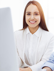 Young cheerful woman sitting at the desk with computer and looking at camera in sunny office. Looks like student girl or business lady. Headshot