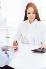 Young business woman and man sitting and working with computer and calculator in sunny office. Bookkeeper checking balance. Tax and audit concepts