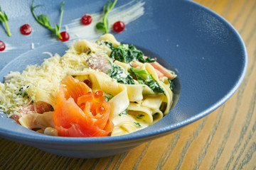Appetizing, home-made tagliatelle pasta with spinach and salmon, parmesan in a blue bowl on a wooden background. Italian cuisine. Add noise on post. Selective focus