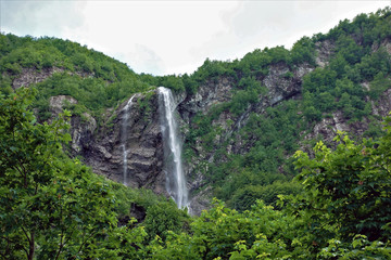 Polikarya waterfall in the Caucasus mountains. Summer. The mountains are covered with green trees. From a great height, a stream of water falls.