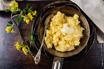 Hot corn porridge with sugar and butter on an old rustic pan and a metal vintage stand. Next to it is a sprig of flowering black currant with yellow flowers