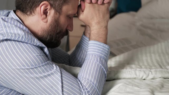 Man is praying. Bearded guy in bedroom in evening with his hands on bed saying prayer