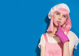 Advertising portrait of housekeeping girl with pink hair that bites a rubber glove on blue background. Hygiene of protection hands. Poster of domestic services of household. Copy space