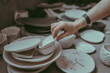 Close-up view of a female hand holding a hand made clay dish amongst other tableware in a workshop. A ceramist in her pottery. A ceramic studio with with her craftswoman. Unique design of dishware.