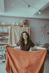 A beautiful smiling young woman with curvy hair standing in the center of ceramic studio with an orange plaid in her hands. Lifestyle shot in a pottery workshop. Ceramist female at work. Cozy interior