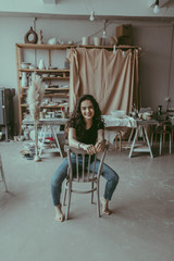 A happy european young woman sitting on a viennese chair in the center of potter studio. A smiling ceramist female at work. Lifestyle shot in a cozy interior of workshop. Clay tableware on background