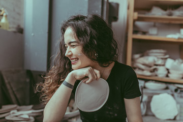 A smiling barefoot young woman in a black t-shirt sitting on a floor with craft clay plate in her hands and surrounded by various tableware and pottery kiln on  background. A ceramist at work concept
