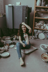 A barefoot young woman sitting on the floor and showing her bare heel to the camera. A ceramist surrounded by various clay tableware. A happy potter girl in her workshop. A lifestyle concept.