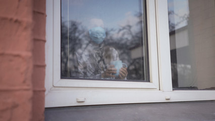 Woman In self isolation Stands near window holding cup Of Tea or coffee at home. Coronavirus pandemic.