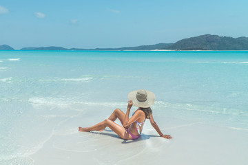 Woman sitting on paradise beach. Tourist on Whitsundays beach, white sand, in pink bikini & hat, with aqua blue turquoise ocean. Travel, holiday, vacation, exotic. Whitsundays Islands, Australia.