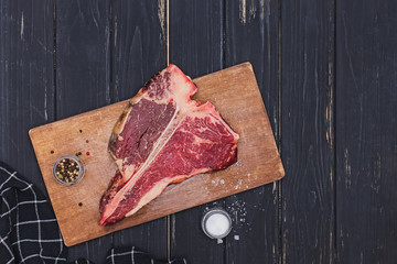Raw T-bone beef steak on the wooden board and spices on black table.
