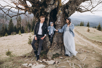 Stylish family in the autumn mountains. Stock photo. A guy in a leather jacket and a young girl in a gray-blue dress with his son stand under a large tree against the sky and mountains