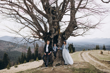 Stylish family in the autumn mountains. Stock photo. A guy in a leather jacket and a young girl in a gray-blue dress with his son stand under a large tree against the sky and mountains