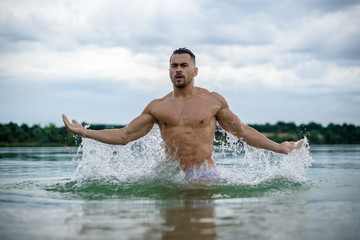 sexy sporty man, macho in white shorts bodybuilder emerges from water, cloudy sky on background.