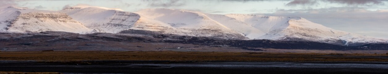 A panorama and wide angle shot of snow capped rocky mountains located on the edge of Hvalfjordur, Iceland. Wispy clouds cover the peaks as the sun rises lighting the mountainside. 