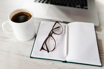 Blank notepad, pen, laptop computer, cup of coffee and glasses on wooden desk. Coffee break, ideas, notes, goals or plan writing concept. Work space. Flat lay office business and finance concept.