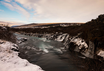 Fototapeta na wymiar A turquoise blue river flows through a snowy autumnal landscape with water cascading over lava rock formations.