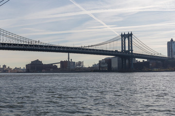 Fototapeta na wymiar Brooklyn bridge seen from afar, side of the bridge, you can see the dark water river and the blue sky, behind the bridge city buildings