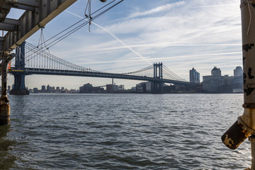 Brooklyn bridge seen from afar, side of the bridge, you can see the dark water river and the blue sky, behind the bridge city buildings