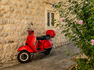 Perast, Montenegro, 20 August 2019. Red Vespa scooter leaning on a white stone wall ith a plant in the foreground.