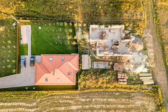 Top down aerial view of two private houses, one under construction with concrete foundament and brick walls and another finished with red tiled roof.