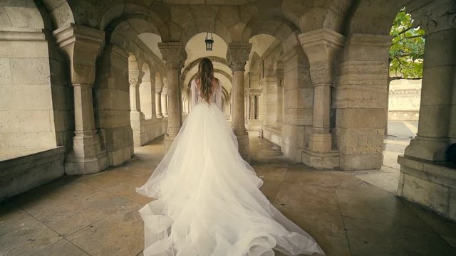 Fashion model, bride in elegant wedding long dress running walks or walks, behind. against the background of the old town and castle architecture, slow motion, columns, arches, historic, buildings