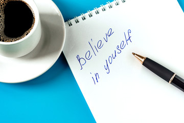 the inscription on the white sheet in the notebook believe in yourself. Black ballpoint pen on white notepad and a cup of coffee on a blue background
