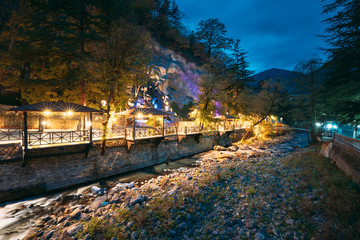 Borjomi, Georgia. Night View Of Borjomula Mountain River Passing Through City, Park And Street Gazebo. Landscape Of Rocky River At Evening Night Illumination.