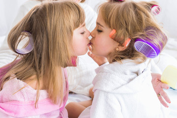 Two daughters are kissing with curlers in hair on the bed in the bedroom. Stay home and family concept.