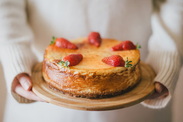 Woman in white sweater standing with fresh homemade cheesecake with strawberries. 