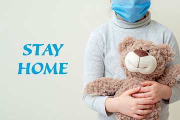 Child is holding Teddy bear wearing protective medical mask with words Stay Home. Health care and virus protection concept.