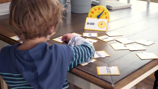 Little Boy Learning Time Using Illustrations And Toy Clock. Panning video showing blond boy sitting in front of the table and he uses cards and toy clock in order to learn the time.