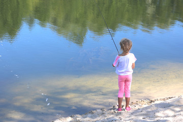 Little child fishing in summer on river on sandy shore against background of blue water and displaying green trees in river