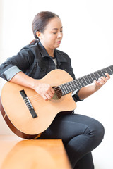 An Asian woman who wears a denim jacket playing a guitar in the house.