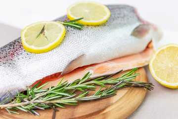A raw decapitated trout sliced on one side with a lemon and rosemary leaves next to it. On a brown wooden chopping board. Freshwater fish.