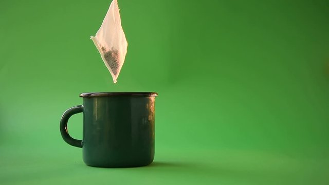 tea-making - a tea bag of 100% cotton on a rope appears on the screen and swings over a green mug with hot water on a green background, steam coming from the mug. the concept of natural tea packs.