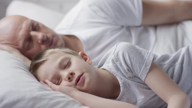 Tracking of bald man in white t-shirt and his peaceful little son sleeping in bed in morning