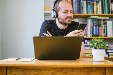 Man working from home office online on computer laptop behind vintage desk with flower in vase, checking smart phone
