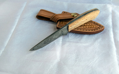 A beautiful wooden handled fixed blade damascus knife displayed on its custom fit leather sheath on top of white fabric portraying elegance with bokeh.