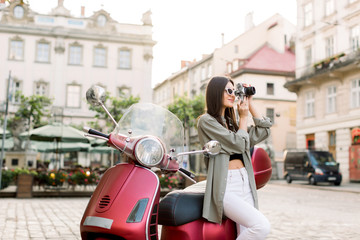 Attractive brunette young woman in stylish clothes and sunglasses making photos of beautiful architecture buildings, leaning on the red scooter on the background of the ancient city street