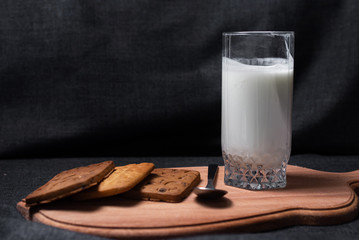 Cookies on a wooden cutting board. A glass of milk and a metal spoon.
Photo on a dark, fabric background