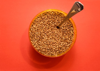 raw buckwheat in a plastic container with a spoon on a red background