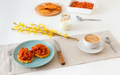Table with homemade spicy baked beans with bacon and potato crumpets, cup of coffee, glass of cream, embellished with few twigs of golden rain. English breakfast.