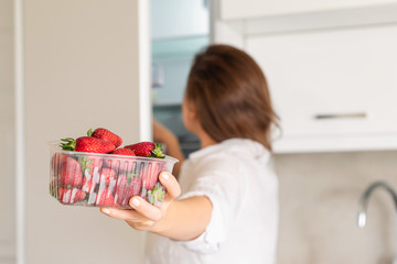 A girl takes out a container of freshly strawberries from the refrigerator. Back view